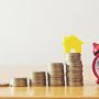 Close-Up Of Coins With Model Homes And Alarm Clock On Wooden Table Against Wall