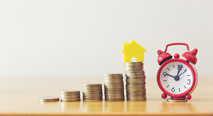 Close-Up Of Coins With Model Homes And Alarm Clock On Wooden Table Against Wall